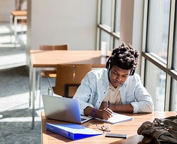 African American teenage boy writes something in a notebook while studying in the campus library. An open laptop is on the table. He is wearing wireless headphones.